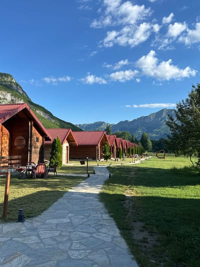 The image depicts a row of identical wooden cabins with red roofs, situated in the city of Gusinje, Montenegro. A stone pathway leads through a manicured lawn, bordered by a line of evergreen trees, towards the cabins. In the background, lush green mountains rise under a clear blue sky dotted with white clouds. To the right of the pathway, a large, red heart-shaped sculpture is visible in the grassy field. The scene suggests a serene and picturesque setting, likely a resort or a tourist accommodation.