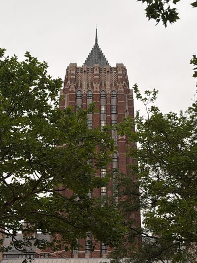This is a photograph of the Sterling Memorial Library at Yale University in New Haven, Connecticut, United States. The gothic-style library tower, constructed primarily of red brick and stone, is partially obscured by lush green trees. The tower features numerous arched windows, some of which reflect the overcast sky, and a distinctive pointed spire at its apex. The image is taken from a low angle, emphasizing the height and grandeur of the building. There are no people visible in the frame.