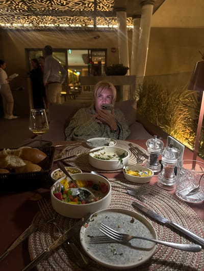 The image shows a woman seated at a table in what appears to be a restaurant or outdoor dining area. The table is set with plates, cutlery, salt and pepper shakers, glasses, and dishes containing food. The woman is holding a phone, seemingly taking a picture. There are other people visible in the background, likely restaurant staff or other patrons. The environment includes decorative lighting and architectural elements, suggesting a modern or upscale setting. The location is potentially El Gouna, Egypt, based on visual style and context clues.
