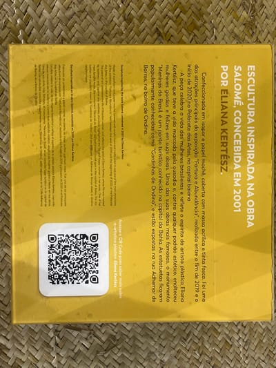 The image displays an overhead view of a yellow, rectangular object, likely a book cover or box, resting on a light brown, woven surface. The object's visible face is yellow, predominantly covered with black text in Portuguese, with some sections translated into English and Spanish. A white square sticker containing a black QR code is affixed to the lower-left portion of the yellow surface.
The primary text, oriented vertically along the right edge, reads "ESCULTURA INSPIRADA NA OBRA SALOMÉ, CONCEBIDA EM 2001 POR ELIANA KERTÉSZ." Further text details an exhibition titled "Fartura e Abundância," which took place at the Palacete das Artes in Salvador, Bahia, Brazil. It references the artist Eliana Kertész's work, including sculptures known as "Meninas do Brasil" and "Cordinhas de Ondina." The description explicitly states that the "Cordinhas de Ondina" sculptures are exhibited on Rua Adhemar de Barros, located in the Ondina neighborhood of Salvador, Bahia, Brazil. Adjacent to the QR code, text in Portuguese instructs the viewer to "Acesse o QR Code para saber mais sobre a artista plástica Eliana Kertész" (Access the QR Code to learn more about the plastic artist Eliana Kertész).
The background consists of a tightly woven material, likely straw or wicker, displaying a diagonal pattern. There are no individuals or discernible actions depicted in the scene.
