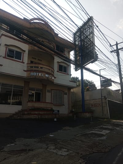 The image shows the exterior of Hotel Wiguna, presumably located in the Zeyathiri District, Myanmar. The building is a multi-story structure with a beige facade. The hotel's name is displayed prominently above the entrance. An advertising billboard stands adjacent to the hotel. Power lines are draped across the sky. The street in front of the hotel is wet and shows signs of wear. A JNE express sign is visible on a wall. A low wall with "Hotel Wiguna" is behind the building and the large sign.