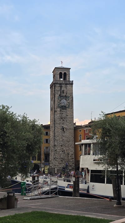 The image captures a vertical view of the Torre Apponale, a prominent clock tower in Riva del Garda, Italy, under a cloudy sky. The stone tower, which is the central focus, has a clock face with Roman numerals and ornate hands. Below the tower, a modern white passenger boat is docked. Several people are visible on the boat's deck and on the adjacent pier. To the left of the tower, a large, leafy tree partially obstructs the view of some buildings. The ground in the foreground is paved with cobblestones and a strip of green grass. The scene suggests a waterfront area, likely a popular tourist spot in Riva del Garda.