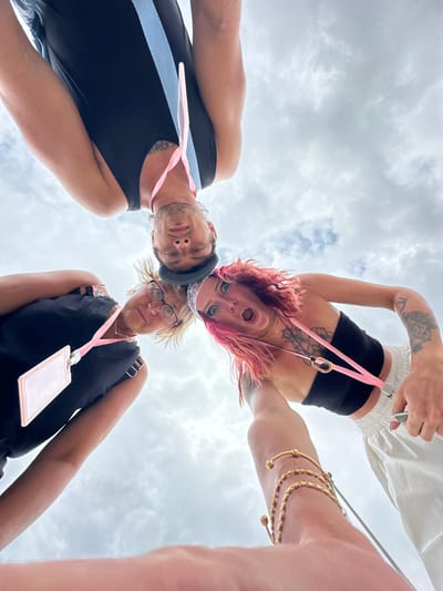 Three people are in the frame, seen from a low angle looking upwards towards a cloudy sky. A man on the left wears a black tank top with a light blue stripe, and a light pink lanyard around his neck. In the middle, a woman with blonde hair and glasses wears a black top with a light pink lanyard holding a badge. On the right, a woman with pink hair and a black top with a light pink lanyard and badge around her neck is looking down at the camera with an open mouth. Her arms are raised; one with gold bracelets. She has tattoos on her arm and chest. All three individuals appear to be looking downwards, towards the camera, with varying expressions. The location is Kampung Toyapakeh, Indonesia.