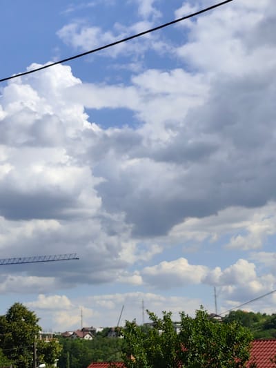 The image depicts a sky with scattered clouds and a black wire crossing the upper portion. Below the sky, a landscape is visible, featuring trees, rooftops of houses, and some distant structures that appear to be power lines and possibly a construction crane on the left. The overall scene suggests an outdoor environment, likely a residential area with some ongoing development, in Durlești, Moldova. There are no visible people or specific actions taking place.