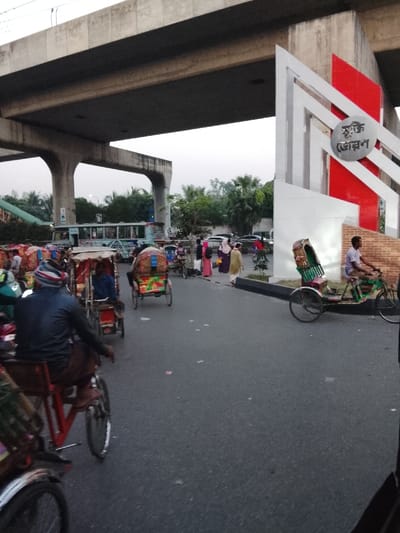 A bustling street scene unfolds under a concrete overpass. Colorful rickshaws dominate the foreground, weaving through the traffic. A bus can be seen further down the road. Pedestrians, some in traditional dress, are crossing near a monument featuring red and white accents and what appear to be Bengali characters. The sky is overcast, contributing to the somewhat muted lighting of the urban environment.