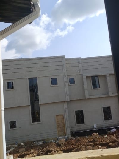 The image shows a section of a light-colored building under construction in Abeere, Nigeria. The building has a modern design with horizontal textured bands on its facade. There are several windows of various sizes and shapes, some are rectangular and dark, reflecting the sky, while one is a tall, narrow vertical window. A wooden door is visible on the lower level. The foreground is filled with disturbed earth, indicating ongoing construction work, with piles of soil, debris, and some building materials like concrete blocks. A white downspout is visible on the left side of the image, attached to an unseen roof. The sky above is partly cloudy with a clear blue sky peeking through. The lighting suggests it is daytime, likely mid-morning or afternoon, with the sun casting a soft light. The overall scene conveys a sense of a developing residential or commercial property in an outdoor setting. No people are visible in the frame. There is no visible text in the image.