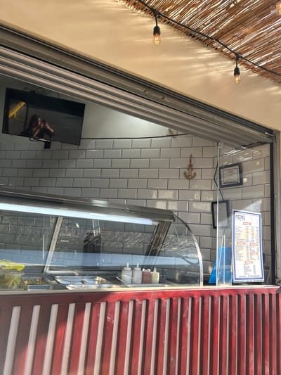 The image shows the interior of a small eatery located at latitude 9.951 and longitude -84.194 in Pozos, Costa Rica. The scene is captured from inside the establishment, looking out towards the front. The front counter is made of red wood with vertical slats. There are several food preparation and display areas behind the counter, including a glass-covered food warmer with a display of various condiments, and a large flat-screen TV mounted to the wall. The walls are tiled in white square tiles. There is a small gold anchor mounted on the wall. A menu is displayed on the right side of the counter. The ceiling is covered in a woven mat of dried reeds. Four incandescent light bulbs are hanging from the ceiling on wires. The image does not show any people, but a person is visible in the reflection of the flat-screen TV.