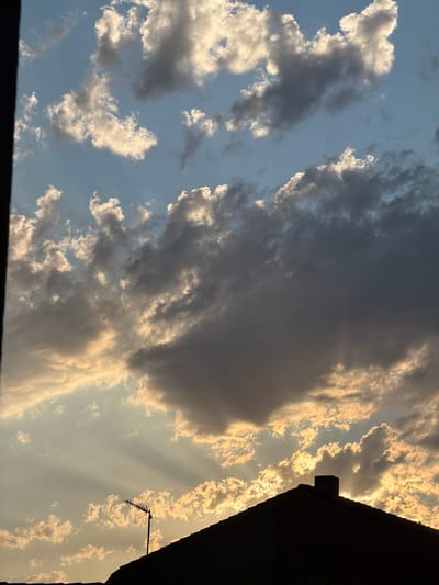 This is a vertical, medium-shot, exterior photograph of a cloudy sky at sunset, viewed from a rooftop or high vantage point, possibly in Arcahueja, Spain. The sky is a gradient of pale blue at the top, transitioning to hues of yellow and orange as it nears the horizon. Cumulus clouds, illuminated by the setting sun, are scattered across the sky, with some appearing white and bright, while others are darker, suggesting they are catching less light or are denser.
In the foreground, silhouetted against the bright sky, is the dark outline of a tiled rooftop with a chimney. To the left of the chimney and slightly forward on the rooftop is a television antenna, its metal structure stark against the warm sky. Sunbeams, or crepuscular rays, emanate from behind a large, dark cloud, fanning out across the sky and illuminating the edges of smaller clouds. The overall impression is one of a dramatic and beautiful sunset over a residential area.