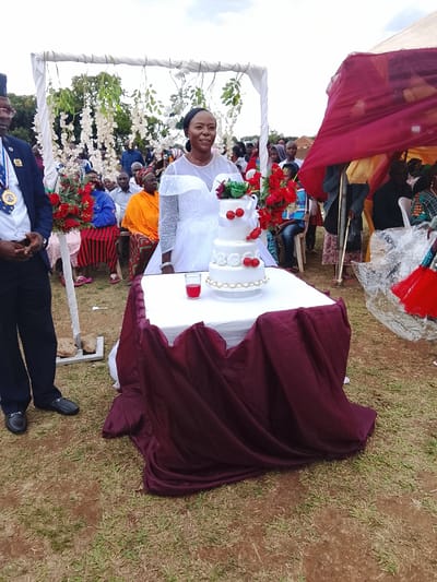 The image depicts a wedding celebration in Kuru, Nigeria. A woman in a white wedding gown stands behind a table covered with a white cloth and burgundy fabric. On the table is a three-tiered white cake decorated with red cherries and white roses, along with a glass of red liquid. To the left of the table stands a man in a blue suit with a medal. Behind the table, an arch decorated with white flowers and green foliage serves as a backdrop. Seated guests are visible behind the arch. To the right of the table, people are standing under a red-draped canopy. The event is taking place outdoors on grassy terrain.