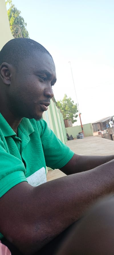 A dark-skinned man in a green collared shirt is captured in a mid-shot. He gazes to the right, with his arms resting in front. In the background, a striped green and white structure stands near greenery and a utility pole. Further back, a building or covered area hosts several people. The sky is bright, indicating daylight. The composition suggests an outdoor setting with the man as the central focus.
