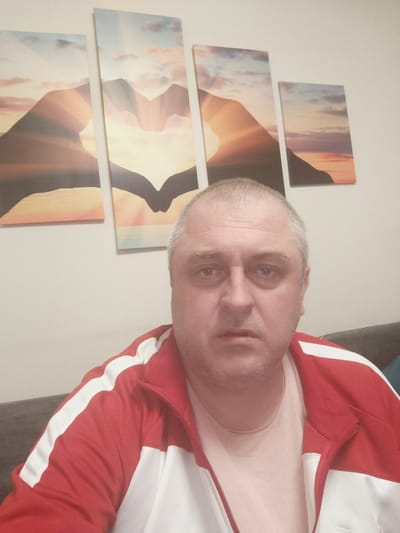 A man is pictured from the chest up, looking directly at the camera. He has short, grey hair, a fair complexion, and is wearing a light pink t-shirt under a red and white zip-up jacket. Behind him, on a light-colored wall, is a four-panel piece of wall art depicting a sunset over mountains, with two hands forming a heart shape in the foreground. The man is seated on what appears to be a dark sofa. The image was taken indoors, with no discernible indication of its specific location within Huntingdonshire, United Kingdom.