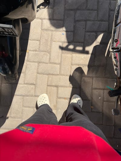 A first-person perspective shot shows a person's legs and feet standing on a paved walkway in Jipang, Indonesia. The ground is made of interlocking gray bricks. To the left, a portion of a black motorcycle is visible, including its rear tire and license plate. To the right, a chrome-plated part of another motorcycle is present, along with a black foot pedal and a green object. A bright red garment, possibly a shirt or jacket, occupies the foreground, obscuring the lower part of the frame. Shadows cast by unseen objects, likely motorcycles and a person, fall across the brickwork. The lighting suggests it is daytime with strong sunlight.