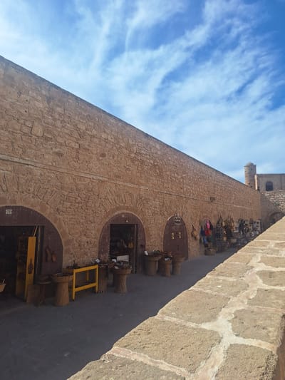 The image depicts a section of the ramparts of Essaouira, Morocco, on a bright, sunny day with a partially cloudy sky. The ramparts are constructed of large, rough-hewn stone blocks, forming a substantial wall that extends from the left of the frame towards the right and into the distance. Along the base of the wall, several arched openings lead into small shops or stalls. These openings reveal goods for sale, including baskets, bags, and other items displayed both inside and outside the shops. One shop has a sign that reads "AZUL SHOP". The ground in front of the shops is paved with dark stone.
To the right of the main wall, the ramparts continue, with a more narrow section of stone-paved walkway or ledge in the foreground. In the background, a defensive tower with a conical roof is visible, characteristic of Essaouira's historical fortifications. The overall impression is one of a well-preserved historical site that is also actively used for commerce. There are no visible people engaged in specific actions, but the presence of the shops implies ongoing activity.