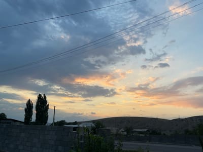 The image depicts an outdoor scene at dusk, with a focus on the sky and a rural landscape. The sky is filled with dramatic clouds, tinged with orange and pink from the setting sun. Several power lines crisscross the upper portion of the frame. In the foreground, a dark, textured wall made of concrete blocks runs horizontally across the lower edge of the image. Behind the wall, two tall, dark green trees stand prominently. Beyond the trees and wall, rolling hills form the background, sparsely dotted with vegetation and what appear to be structures. A road is visible in the lower right quadrant. The overall impression is one of a tranquil, rural evening. The location is identified as the city of Taran-Bazar, Kyrgyzstan. There are no people visible in the image.