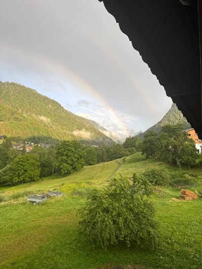 This image captures a serene landscape in Saint-Jean-d'Aulps, France, featuring a double rainbow arcing across a cloudy sky over lush green mountains. In the foreground, a grassy meadow slopes downwards, dotted with trees and a few hay bales. To the left, two trailers are parked in the tall grass, with a winding path leading up the hillside. In the mid-ground, a village of wooden chalets nestles amongst the trees at the base of a steep, forest-covered mountain. The sky is overcast, with patches of sunlight breaking through, illuminating sections of the mountainside and creating a misty atmosphere. The scene is framed by the dark, wooden eaves of a building in the upper right corner, suggesting a view from a balcony or covered porch.