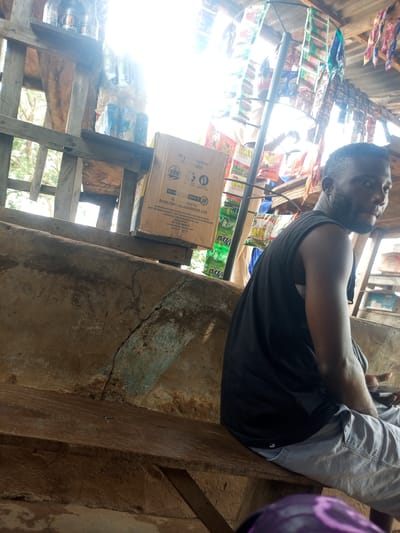 In Iyara, Nigeria, a Black man wearing a black tank top and grey pants is seated on a wooden bench. He holds a mobile phone in his right hand, his thumb resting on the screen. The bench is positioned in front of a concrete structure that appears to be a wall or stall. Behind the man, a shelf holds various bottled drinks, including a large bottle with a label showing a smiling face. Above the shelf, a rack displays an assortment of colorful packets. A cardboard box labeled "SUNLIGHT HYGIENICS LTD." is also visible on the shelf. The setting suggests an outdoor market or a small shop, with natural light filtering in from above.