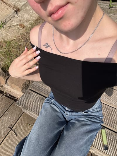 The media file shows a close-up of a person lying on a wooden deck outdoors. The person is wearing a black tank top and blue jeans. A silver necklace with a pendant is visible around their neck. Their nails are painted white with glitter. A small tattoo of a leaf branch is on their collarbone. The setting appears to be a sunny day, with strong shadows cast on the deck. Dried grass and some rocks are visible around the deck. A green vape pen is lying on the deck next to the person's leg. The text "VAPORESSO" is visible on the vape pen. The scene suggests a relaxed, casual moment outdoors in Pozdyshki, Russia.