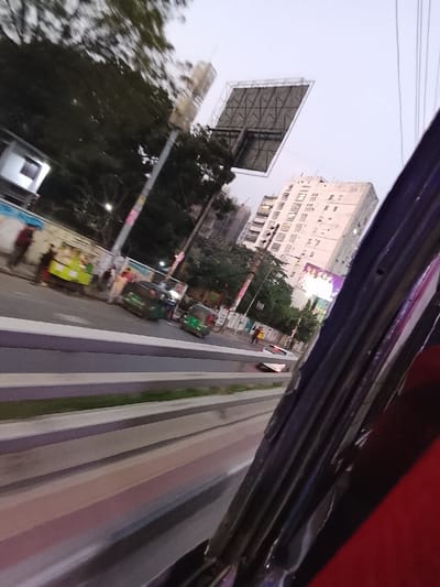 A street view, likely taken from inside a vehicle. Visible are buildings, trees, and an advertisement billboard. Traffic includes green auto-rickshaws. Barriers line the road. The image has a tilted perspective and a part of the vehicle frame is visible on the right side. The lighting suggests either early morning or late afternoon.
