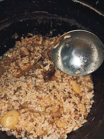 In Кош-Дөбө, Kyrgyzstan, a close-up, overhead view shows a black pot containing a cooked rice dish. The dish appears to be a pilaf or similar rice preparation with visible grains of rice, small green peas, pieces of meat, and slices of potato. The food is in a liquid broth, and some of the rice grains appear slightly clumpy, suggesting it has been recently cooked or is being served. A large, silver ladle rests on the right side of the pot, partially submerged in the food, with its handle extending out of frame. The pot itself is dark and appears to be made of cast iron, with some residue visible on its inner rim. The lighting is dim, and the overall impression is of a rustic, home-cooked meal.