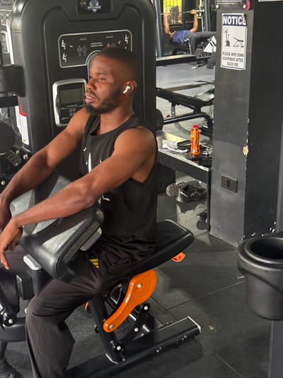 A Black man with short hair and a beard is sitting on a black and orange gym machine in a gym. He is wearing a black sleeveless t-shirt and black pants. He has white earbuds in his ears and is looking to his left. In the background, another person is seen on a bench press, and a "Notice" sign is posted on a black column. The gym floor is dark grey, and there are other gym equipment visible in the background. The environment appears to be an indoor gym.