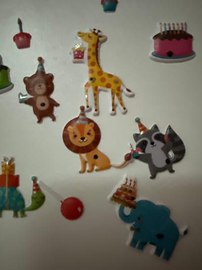 This media file contains a collection of colorful, cartoon-style stickers or decorations adhered to a white surface, likely a wall. The subjects are primarily cartoon animals and celebratory items, suggesting a birthday or party theme.
The visible objects include:
* A pink birthday cake with candles.
* Two raccoons, one appearing to be on top of the other, both wearing party hats.
* A blue elephant with a party hat.
* A yellow giraffe with orange spots, also with a party hat.
* A lion with a party hat.
* A brown bear wearing a party hat and a vest.
* A red lollipop on a stick.
* A green creature, possibly a duck or frog, wearing a party hat and a patterned garment.
* Several gift boxes.
The activity or event implied is a birthday celebration. The presence of party hats, cake, and gift boxes strongly supports this interpretation. The cheerful and playful nature of the cartoon characters evokes a celebratory and joyful atmosphere.
There are no explicit time of day or weather cues. The background is a plain white surface, offering no specific location context beyond what can be inferred from the theme of the stickers. There is no visible text within the media file. The context of Getafe, Spain, is provided externally and is not discernible from the image itself.