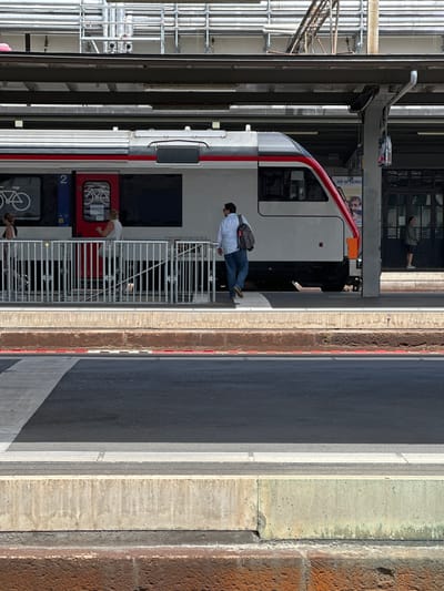The image depicts a train station platform in Lausanne, Switzerland. In the foreground, a dark asphalt surface with white directional lines and tactile paving is visible. A concrete barrier separates this area from a raised platform. The platform itself features a train, specifically car number 2, which is white with a red stripe and a red-nosed front. A man in a white shirt and blue jeans, carrying a backpack, walks away from the train. Two women are also on the platform near the train entrance, one looking towards the train and the other walking away. In the background, the station's covered roof structure, with exposed piping and scaffolding, is visible. Further back, a building facade with large glass doors and a poster with images of people can be seen. Another man in a green shirt stands near the building entrance. The overall scene suggests a typical day at a European train station.