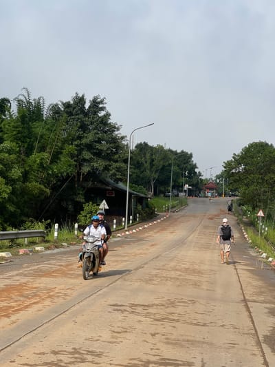 A man and a boy on a scooter drive down a wet, dirt-covered concrete road lined with trees and bamboo. The man, wearing a white shirt and blue helmet, drives the scooter while the boy, wearing a black shirt, sits behind him. A person with a black backpack walks away from the camera on the right side of the road. In the distance, a red-roofed building with a gate is visible. The sky is overcast.