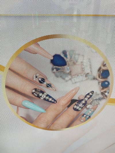 The image displays a close-up view of two hands with elaborately decorated fingernails, possibly from a nail salon advertisement or display. The nails are adorned with various designs, including dark blue polish with white accents, intricate patterns with rhinestones, and solid light blue polish. The hands appear to be resting or posing, and the overall impression is one of detailed nail art. The image is overlaid with a perforated pattern, suggesting it might be a print on a screen or a similar textured surface. There is no discernible location in the city of Rosenheim, Germany, visible in this image.