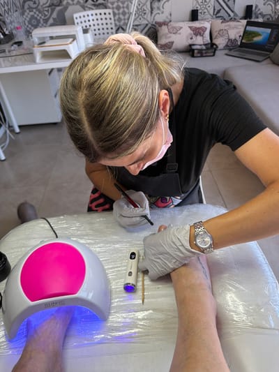 A woman, wearing a black shirt and white gloves, is performing a pedicure on a person's foot. She is leaning over the foot, which is placed inside a white and pink LED nail lamp. The lamp is emitting a blue light, indicating it is in use. The woman holds a red-tipped tool in her gloved hand, appearing to be applying something to the toenails. A silver wristwatch with Roman numerals is visible on her left wrist. In the background, a laptop displays an image of a landscape. The scene takes place in a brightly lit room, possibly a nail salon, with tiled walls and furniture that suggests a professional setting. A small device with a blue light is also present near the nail lamp. The overall atmosphere is focused and professional.