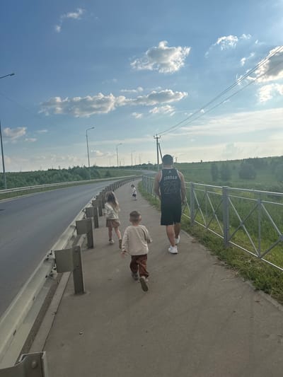 A group of four people, an adult male and three children, are walking along a paved sidewalk next to a road. The adult male, wearing a black tank top with "JORDAN 23" on the back and black shorts, is walking away from the camera. He is wearing white sneakers. To his left and slightly ahead are three children. The closest child is a boy wearing a light-colored long-sleeved shirt and brown pants, also walking away from the camera. To his left is a girl with long blonde hair, wearing a light-colored jacket and a skirt, walking in the same direction. Further ahead and to the right is another young child, partially obscured, wearing a white t-shirt and shorts. They are walking along a sidewalk bordered by a metal guardrail and a grassy embankment with trees and foliage beyond. The road to the left is empty. Overhead, the sky is blue with scattered white clouds, and power lines are visible. Streetlights are positioned along the side of the road. The scene appears to be in a semi-rural or suburban area.