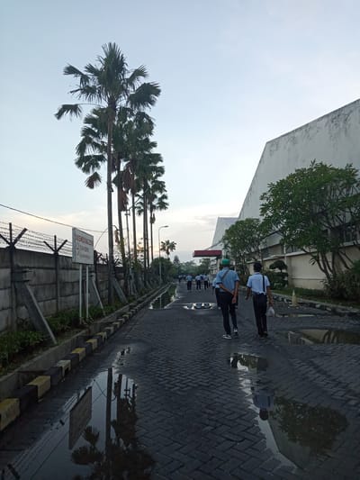 The image captures an outdoor scene on a paved road, likely within an industrial or institutional area in Sukomoro, Indonesia, during either early morning or late afternoon. A group of men, appearing to be workers, walk away from the camera down the road. Most are wearing light-colored shirts and dark trousers; one man in the foreground wears a green cap.
The road is dark gray, made of interlocking paving stones, and features numerous puddles reflecting the sky, trees, and structures. The puddles suggest recent rain or standing water. Yellow and black painted curb blocks line the road's edge.
To the left, a tall, gray concrete wall topped with barbed wire runs alongside the road. Behind this wall and further along the path are several tall palm trees. A white rectangular sign on a pole is visible near the wall, stating "NO SMOKING ZONE" and "KAWASAN DILARANG MEROKOK," which translates to "No Smoking Area" in Indonesian.
On the right side of the road, a large, light-colored building with a sloped roof stands. Its facade is partially screened by green trees and shrubs. The sky is pale blue with a soft, warm glow near the horizon, indicating a low sun angle. The overall scene shows people commuting along a functional pathway in a semi-industrial setting.