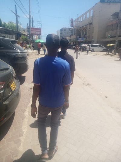 The image depicts an outdoor street scene under clear daylight, identified as Shanzu ward, Kenya. The perspective is from behind two male pedestrians walking forward on a paved sidewalk.
The closest individual, a male, is wearing a blue short-sleeved t-shirt with thin red and white trim on the sleeves, dark slim-fit trousers, and dark sandals. He has a dark baseball cap on, worn backward or covering the back of his head. A light blue wristband is visible on his right wrist. Directly behind him, a second male is partially visible, wearing a dark short-sleeved t-shirt and dark trousers.
To the immediate left of the foreground individuals, two dark-colored cars are parked on the paved sidewalk. The visible license plate on the closer car reads "KCV 843P". The sidewalk features light-colored rectangular paving blocks. Further down the street on the left, a red sign with white lettering displaying "BUTCHERY" is visible above street-level activity and buildings. Numerous overhead utility lines crisscross the sky.
To the right of the foreground individuals, the paved sidewalk transitions to a dirt or gravel shoulder, then a gray asphalt road surface. Multiple pedestrians are visible walking along the street and distant sidewalks. A white van is parked on the street further down, and a motorized tricycle with a yellow canopy is also present. Multi-story buildings line the right side of the street, one bearing a "CO-OPERATIVE" sign at its top, and another featuring a vertical blue sign with "nai as" lettering. The overall scene suggests a busy commercial or residential street environment.