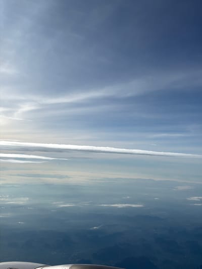 An aerial view shows the sky above Laem Sak, Thailand. The sky is a gradient of blue, with wispy white clouds stretching horizontally across the frame at various altitudes. Below the clouds, a hazy landscape of mountains and mist is visible, suggesting a high vantage point, likely from an airplane. The lower portion of the image shows the edge of an aircraft engine cowling, confirming the aerial perspective. The scene is devoid of any people or specific urban structures.