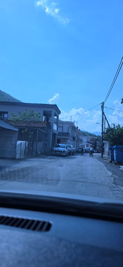 The image displays a street scene in Melivoia, Greece, captured from inside a vehicle, with the dashboard visible in the lower foreground. The time is daylight, under a clear blue sky with scattered cumulus clouds. The street is paved, exhibiting visible wear and patching. Two-story residential buildings with light-colored facades line both sides of the street, some featuring balconies and roof-mounted satellite dishes. A white Peugeot SUV, bearing Greek license plate YAN-5077, is parked on the left side of the street, followed by a lighter-colored pickup truck. In the middle distance, two individuals are riding bicycles away from the viewer down the street. On the right, a utility pole supports numerous overhead power lines, and two blue municipal waste bins are positioned on the sidewalk. Vegetated mountains form the background, indicating the town's geographic setting.