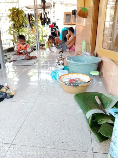 The image shows an outdoor patio in Gegesik, Indonesia. Three people are visible. One is an adult male sitting on the floor with his legs crossed, looking towards the right, and another is a young boy sitting on the floor with his legs crossed, looking down at a small, rectangular object. There is also a young girl wearing a pink shirt sitting on the floor, looking to the left. The adult male is wearing a dark gray t-shirt and the boy is wearing an orange and white shirt with a blue stripe.  The girl is wearing a pink shirt and black pants. The patio has a light gray tiled floor, and there are numerous objects scattered around, including a small metal teapot, a blue plastic basin, a large wicker plate, and a stack of banana leaves.  The banana leaves are next to a blue and white bucket. There are several other objects visible, including a white plastic bag, a white water bottle, and a knife on the banana leaves.  The patio is situated next to a building that has a brown wall.  The building has several large windows.  A large green leafy plant is visible to the left of the patio, which may be part of a garden.  The image was taken at 07:54 on April 9, 2025.  There are several other objects in the image, including a white water bottle, a white plastic bag, and a knife.