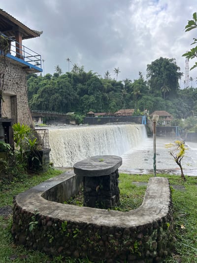 The image captures a wide, cascading waterfall in a lush, green environment. The waterfall flows over a constructed dam, with a strong current evident in the churning white water. On the right side of the frame, a curved concrete structure, possibly a bench or part of a decorative element, is visible, bordered by dark stones and overgrown with moss and small plants.

In the background, dense, tropical vegetation dominates the landscape, with tall trees and palm trees reaching towards the overcast sky. Several small, traditional-style buildings with thatched roofs are nestled amongst the trees near the top of the dam, suggesting a resort or tourist area. A metal railing and blue stairs are also visible along the edge of the dam, indicating pathways for visitors.

On the left side of the image, a stone building with a pitched roof is partially visible, topped with a blue metal railing. A telecommunications tower can be seen in the distance, rising above the trees. The overall scene is characterized by a natural, yet developed, setting, likely a popular spot for recreation or sightseeing.

The weather appears to be cloudy, contributing to the diffused lighting and slightly muted colors of the scene. No people are visible in the image, and no text is readily apparent. The time of day is not clearly indicated but seems to be daytime, given the ambient light. The atmosphere suggests a tranquil, somewhat humid tropical day. The location context provided is Klungkung, Indonesia, which aligns with the tropical vegetation and architectural style.