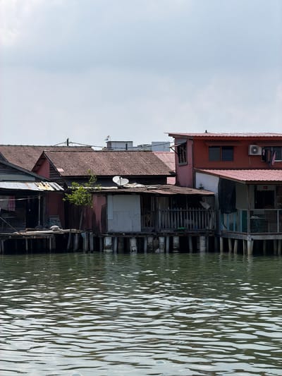 This image depicts stilt houses along the waterfront in George Town, Malaysia. The houses are constructed from wood and metal with a mix of tile, corrugated and painted roofs. The houses are positioned directly above the water, with wooden supports extending into the water. A small tree has grown up between the houses. Laundry is hanging on clotheslines. The water has a distinctive wave pattern. The sky is overcast.