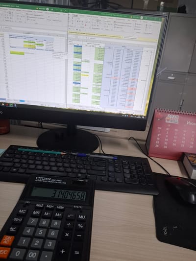 The image depicts a desk scene, likely in an office or work area. The primary focus is a computer monitor displaying an Excel spreadsheet with financial data. To the right of the monitor is a red desk calendar for June 2026. In front of the monitor sits a black keyboard and a black computer mouse. Below the keyboard, a black CITIZEN SDC-444S calculator is visible, displaying the number "31404658". The environment appears to be a workspace, with the lighting suggesting it might be indoors during the day or under artificial light. There are no people visible in the image. The presence of a calendar and financial spreadsheet suggests a professional or business context. The location is not specified in the image, but if it were in the city of Tecnimont camp, Russia, it would imply a remote or industrial work setting.