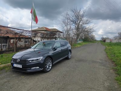 A dark gray Volkswagen Passat Variant is parked on a dirt road. The car faces the viewer, with its front passenger side angled slightly towards the camera. Behind the car, to the left, is a weathered structure with a red tiled roof and a leaning wooden framework. The Bulgarian flag flies from a pole behind this structure. To the right of the car, the dirt road stretches into the distance, bordered by green grass and sparse vegetation. A bare tree stands next to the road on the right. The sky is overcast with dark clouds. The license plate on the car reads "A 2203 PP." The scene appears to be a rural or semi-rural area within Krushovo, Bulgaria.