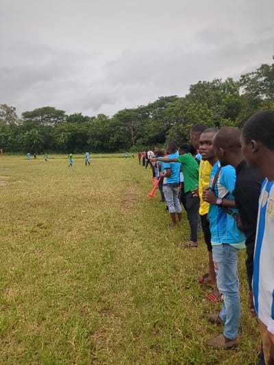 A group of young men stand in a line along the edge of a grassy field, watching a soccer game in progress. The field is surrounded by trees. The sky is overcast. The game is being played by a group of young men, wearing blue and green jerseys. The men in the line are wearing various clothing, including blue and green shirts, and one is holding a red plastic horn. The scene takes place in a field on the outskirts of the city of Morogoro, Tanzania, at coordinates -6.799, 37.63.