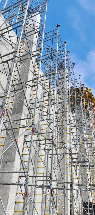 The image depicts a construction site with a large concrete structure and extensive scaffolding against a blue sky with scattered clouds, suggesting it's daytime. The scaffolding is made of metal poles and crossbars, with yellow rungs forming ladders. The concrete structure appears to be a tall column or wall, typical of large-scale construction projects. The scene is a close-up view, emphasizing the intricate network of scaffolding and the raw, unfinished concrete. The overall impression is one of ongoing construction and industrial activity. There are no people visible in the frame. Some scaffolding components have markings, for example, a label with the text "100" is visible on some horizontal poles. The location is Primorye, Russia.