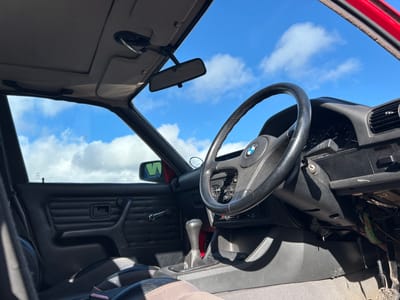 The image shows the interior of a car, specifically the driver's side dashboard, steering wheel, and front window, with a view of a bright blue sky with white clouds visible through the windshield. The car appears to be in a state of partial disassembly, with exposed wiring under the dashboard. The rearview mirror is visible. The car's interior is a mix of black and light brown/beige. The location in the city of Rutherglen, United Kingdom cannot be determined from the given image.