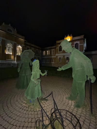 In the city of Kislovodsk, Russia, illuminated by night, a scene unfolds featuring topiary sculptures set against the backdrop of a building. Three green, bush-like figures depict a family: a man in a hat holding a cane, a woman with an umbrella, and a young girl holding a bouquet. The figures are positioned on a circular cobblestone patio. A wrought-iron carriage-like object is also present. The building behind them exhibits illuminated windows and architectural details, suggesting it is a historical or cultural site.
