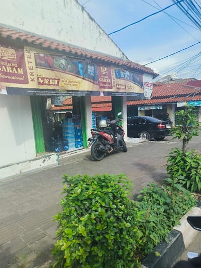 The scene shows the front of a small store in Purwakarta, Indonesia, located at coordinates -6.545 latitude and 107.442 longitude. The store has a white facade, a brown tiled roof, and a large sign advertising "Gudang Garam International" and "Kreteks" with a price of Rp. 18,000. The sign is attached to the front of the store, which appears to sell beverages and other products. A red motorcycle is parked in front of the store. A black car is parked on the side of the street in front of the store. The store's windows are covered with green, slatted blinds. There are several green plants in the foreground of the scene. A small section of the street is visible in the foreground. The sky is clear blue, with some wispy clouds. The street and sidewalk are paved with grey concrete blocks. Power lines run across the top of the image.