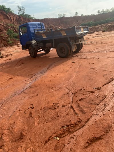 A blue dump truck is parked on a red dirt surface in what appears to be a quarry or construction site. The truck has a flatbed and dual rear wheels. The surrounding environment consists of reddish-brown soil banks and sparse vegetation. The sky is partly cloudy. The scene suggests an active or recently used industrial area, possibly in or around Ijesha-Ijebu, Nigeria.