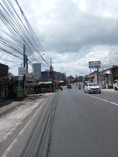 The image depicts an urban street scene in Gubbisamamidi, India, under an overcast sky. The multi-lane asphalt road features a solid white line on its left edge and a dashed white centerline. Shadows from overhead wires are cast prominently across the left lane.
**Traffic and Objects:** A white compact car travels in the right lane, heading away from the viewer. Behind it, a dark-colored sedan follows. Multiple motorcycles are present on the road, with at least four visible, interspersed among the cars and moving in various directions, primarily away from the viewer. A portion of a white utility truck is visible on the far right.
**Left Roadside:** The left side of the road features a concrete sidewalk bordered by a row of single-story commercial structures, some with corrugated metal roofs. Prominent signage includes "SHOP Bisa" and a "Canon" logo. A dense network of black utility wires is strung between numerous dark utility poles along this side of the street. Two individuals are visible near the shops; one person in light-colored clothing and a head covering stands near an entrance.
**Right Roadside:** On the right, a large red, blue, and yellow "Indomaret" sign is prominently displayed, indicating a convenience store, a chain typically found in Indonesia. Behind this sign, a petrol station canopy is partially visible. Other commercial buildings line this side of the street. Green foliage is visible in the background and behind some structures.