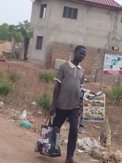 A man in a gray polo shirt and dark pants walks on a dirt road near Dungu, Ghana. He carries a shopping bag with "Aqual" printed on it. A two-story, unfinished concrete building stands in the background. A sign with images is positioned next to him. The surrounding area is dry and littered with debris.