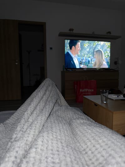 The image shows a dimly lit room with a television playing a scene with a man and a woman in an outdoor setting. The TV is mounted on a wall above a wooden cabinet. Next to the TV is a wooden bedside table with a glass of water and a HalfPrice shopping bag on it. A textured, light-colored blanket or throw is draped over something in the foreground, obscuring much of the right side of the image. The location, Balatonlelle, Hungary, cannot be verified from the given image.