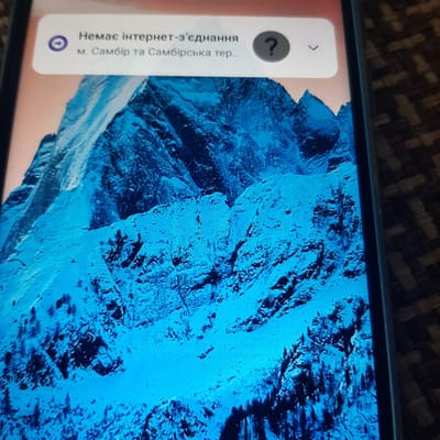 The image shows a close-up of a smartphone screen displaying a notification. The background of the phone screen features a vibrant blue, abstract image that resembles a rocky, mountainous landscape. The notification is in the lower-left corner of the screen, presented within a pale pink rectangular box. The notification text is in Ukrainian and reads: "Немає інтернет-з'єднання" which translates to "No internet connection". Below this, it says "м. Самбір та Самбірська тер..", indicating the location as "Sambir city and Sambir district..". There is also a circular icon with a question mark within it next to the text, and another circular icon with a Wi-Fi symbol crossed out below the question mark icon, further emphasizing the lack of internet connection. The overall scene suggests a user troubleshooting an internet connectivity issue on their mobile device. The lighting appears to be artificial, consistent with indoor use of a phone.
