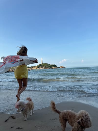 A woman with windblown hair, wearing a white patterned kimono and yellow shorts, stands on a sandy beach facing a lighthouse on a rocky island. Two dogs, one light brown with pink dyed fur and the other a tan poodle, are on the beach near her. Gentle waves lap the shore under a clear blue sky. The scene appears to be at Mũi Kê Gà, a peninsula in Thuận Thành, Vietnam.